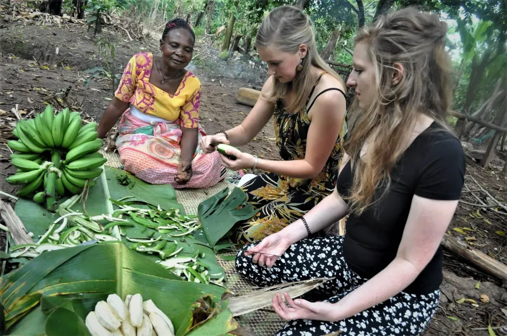 Photo of a Ugandan woman seated in the garden peeling bananas with two white tourists